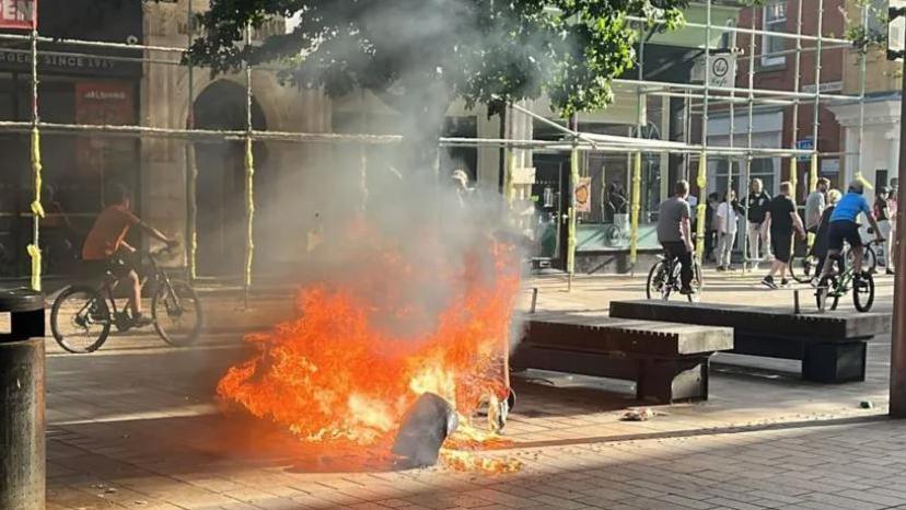 A bin on fire in Hull city centre. In the background, boys ride by on bikes towards a small crowd of people at a street corner