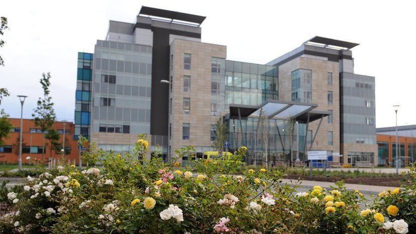 The outside of Peterborough City Hospital, with a big glass atrium. In the foreground are flowers.