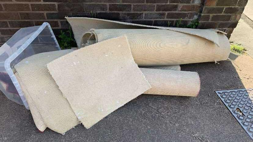 White rugs rolled up on the pavement. There is a plastic box on its side with off-cuts of carpet in it.