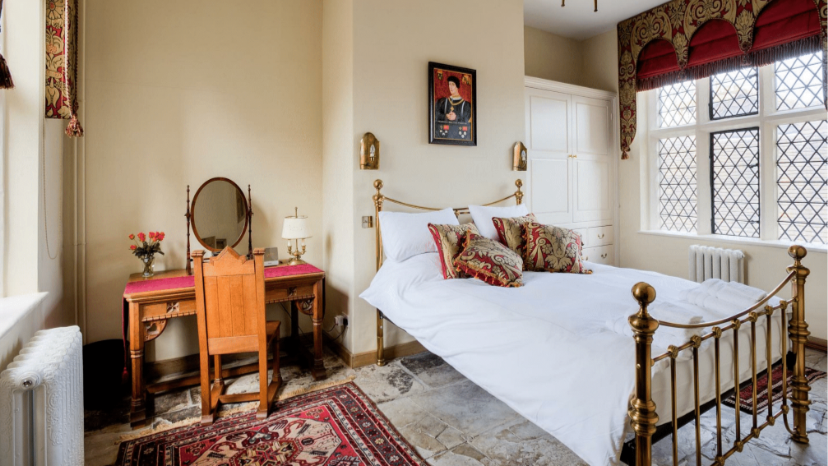 A room with a gold four poster bed, and a portrait hanging above it. A large window on the right has latticed markings on it, as well as a royal red curtain with red and gold decor. There is a brown wooden dressing table and chair, with a red runner on the table, and a mirror on it.