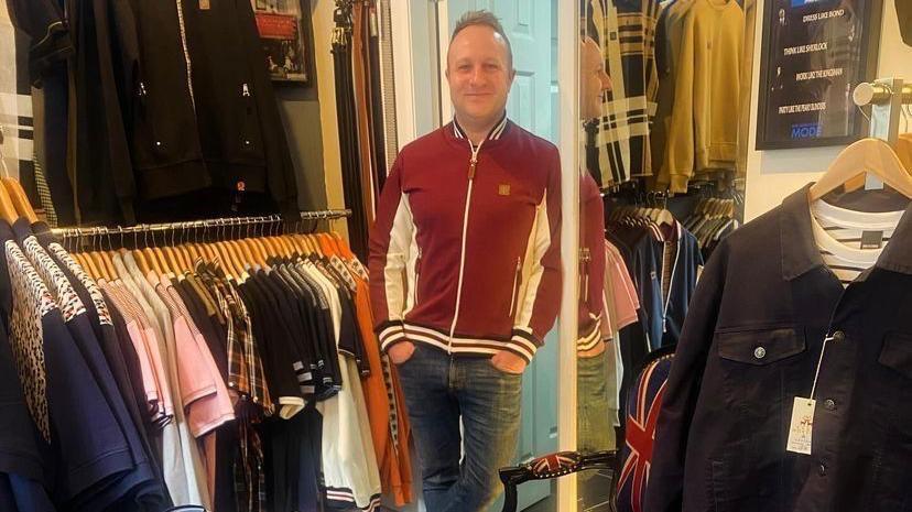 a man wearing a red zip up hoodie and jeans is leaning against a full length mirror in a clothes shop.