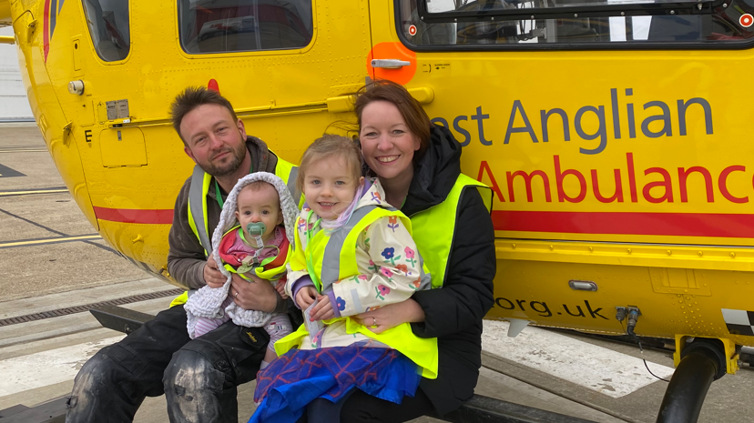 Russ Whitehouse, Claire Whitehouse, both in hi-vis waistcoats, sitting on the footboard of a yellow East Anglian Air Ambulance helicopter, while holding their young daughters on their laps 