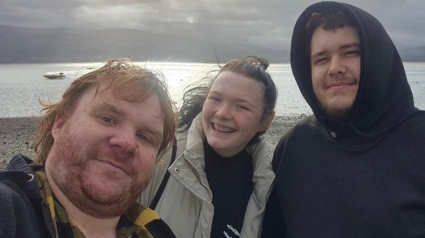 Three friends posing for a selfie on a beach with a boat on the water behind them. They are wrapped up warm, one of them has a hood up, and it looks a cloudy day although the sun is shining through onto the water.