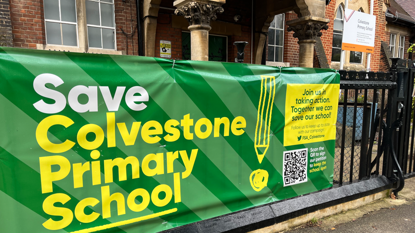 A black iron railing in front of Colvestone Primary School with a green and yellow tarpaulin banner reading 'save Colvestone Primary School'