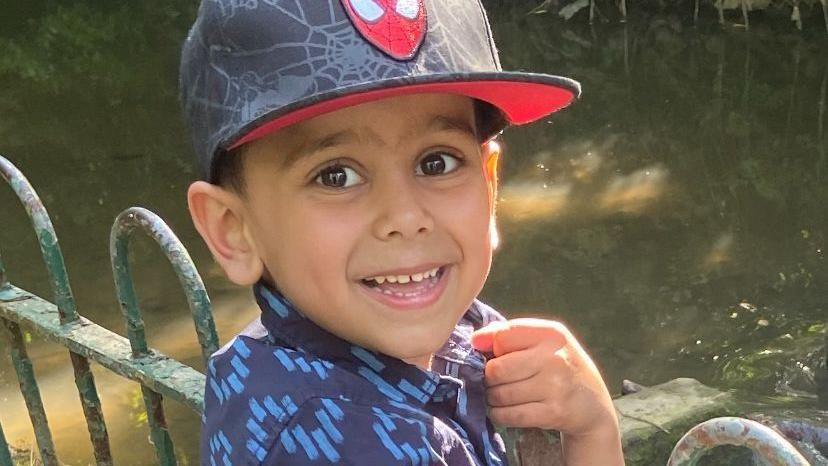 Yusuf Nazir, a small boy, stands next to some railings by a stream. He smiles for the camera and wears a blue short sleeved shirt and a baseball cap with a Spider-Man logo on it.