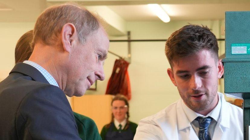 A side-on view of the Duke of Edinburgh in a dark suit talking to a young man wearing a white lab coat and tie