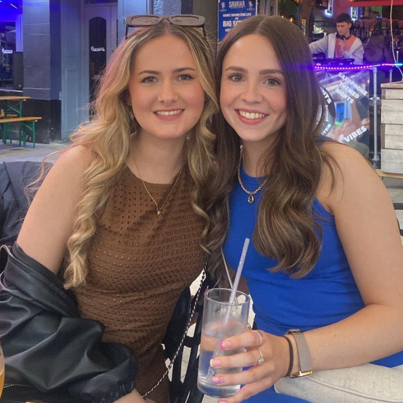 Cerys Davage, with blonde hair and brown dress, with a friend, who is dark haired and wearing a blue dress and holding a drink. They are in a bar and smiling at the camera