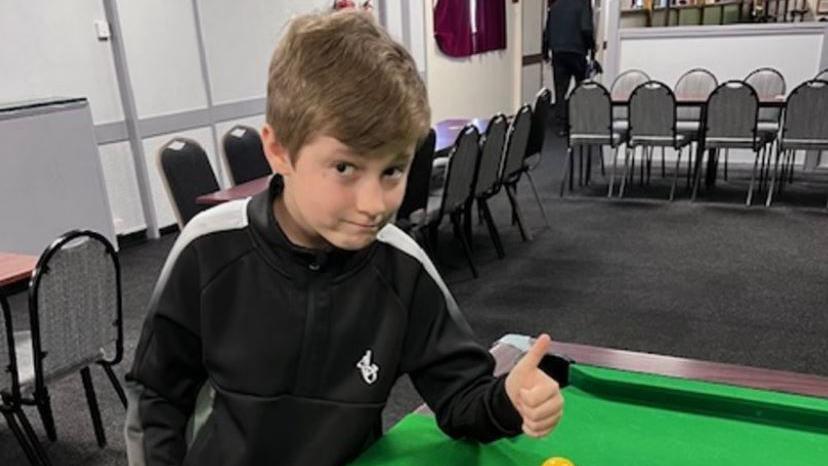 A boy in a black and white tracksuit giving a thumbs up signal as he stands over a pool table, with the balls racked up in a triangle. He is in an events room, which has chairs around the outside. The boy has dark brown hair and is half-smiling.