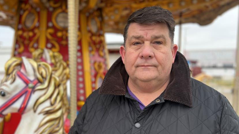 Derek Gray, a man with short black hair who is looking at the camera and is wearing a black coat and purple top. He is standing in front of a golden carousel and carousel horse.