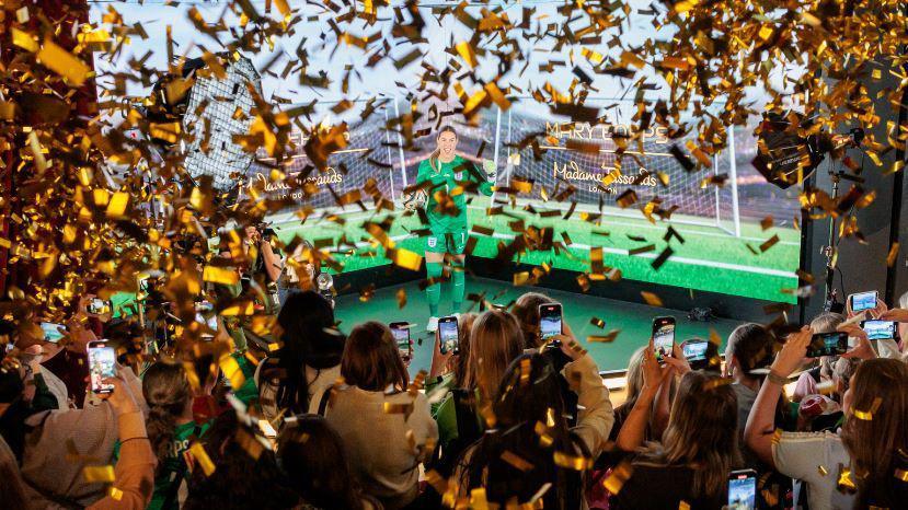 A number of fans hold their phones towards a waxwork of Mary Earps in her green England kit. There is gold confetti in the air. 