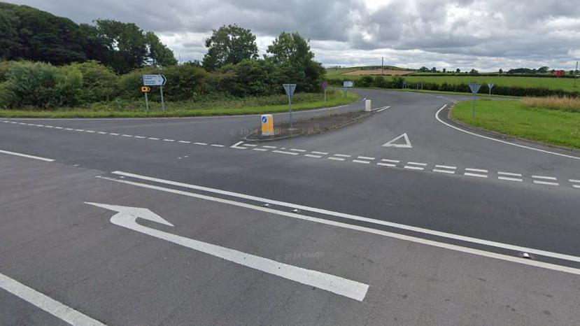 Streetview of a junction, with a large white arrow in a turning lane in the middle of the road pointing towards a road heading off round a corner. There is a yellow and white traffic cone at the mouth of the junction and a sign pointing up the road. Green trees and fields are beyond.