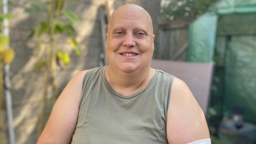 Faye Woodley looking ahead and smiling. She is sat down in a sunny garden. She has no hair and is wearing a light green tank top.