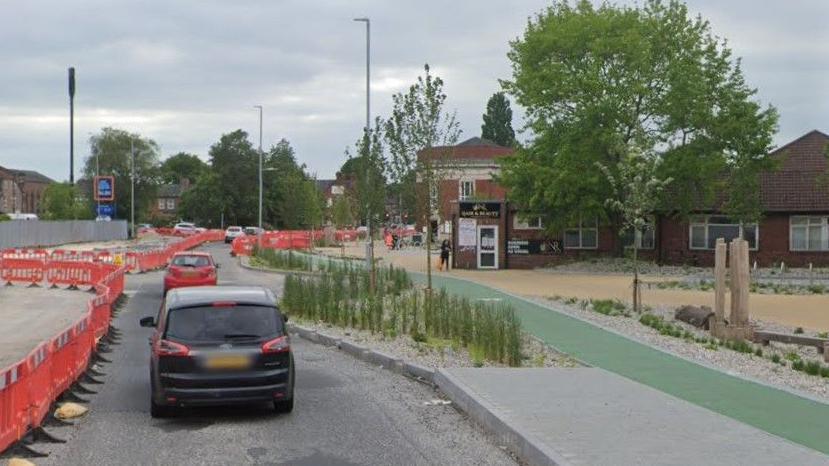 Two cars drive on a single-carriageway road alongside orange panels on the boundary of roadworks and a green cycle path, which runs adjacent to yellow paving and green trees and bushes