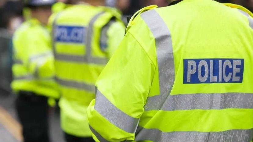 The back of an unidentifiable police officer wearing a high-visibility jacket saying 'police' on it, with two more officers in soft focus behind him.