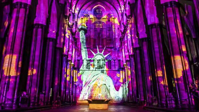 Luxmuralis installation which shows the statue of liberty on the back of the cathedral. Purple and orange lights flank the image.