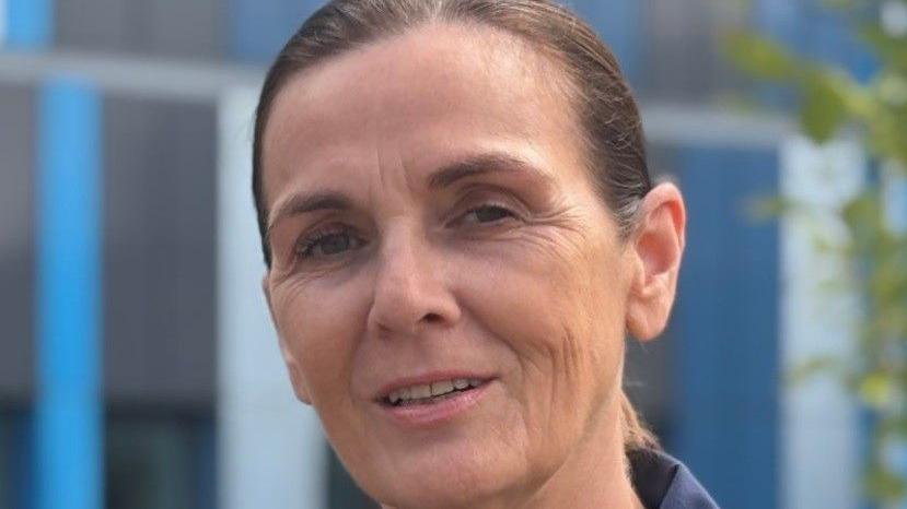 A woman faces the camera. She is wearing a navy blue shirt with yellow trim around the collar and has her hair tied behind her head.