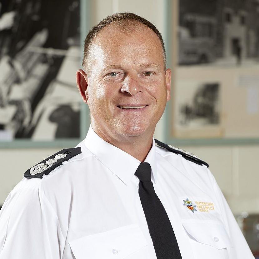 A portrait photograph of chief fire officer Phil Shillito. He has short cropped hair and is smiling into the camera. He is wearing a white shirt with a Humberside Fire and Rescue logo, black lapel badges and a black tie.