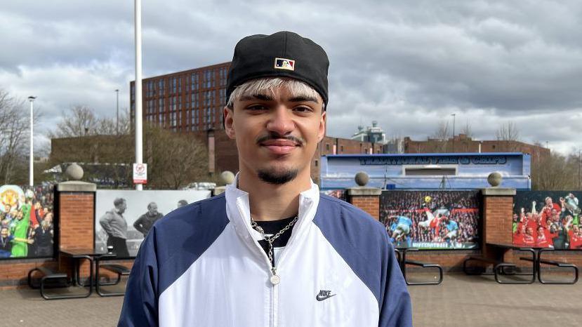 Joao with blond hair and black beard wearing a black cap white and blue sports jacket with silver necklace. 