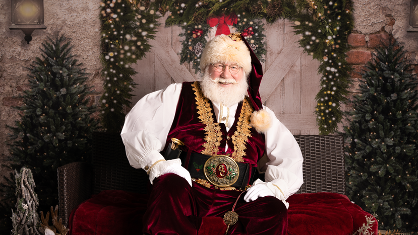 Allan Evans, in a full Santa suit, sat on a velvet red throw with Christmas trees in background and woodland animal ornaments at his feet