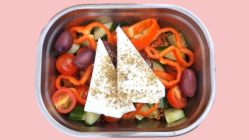 A Greek-style salad in a tin lunchbox on a pale pink background