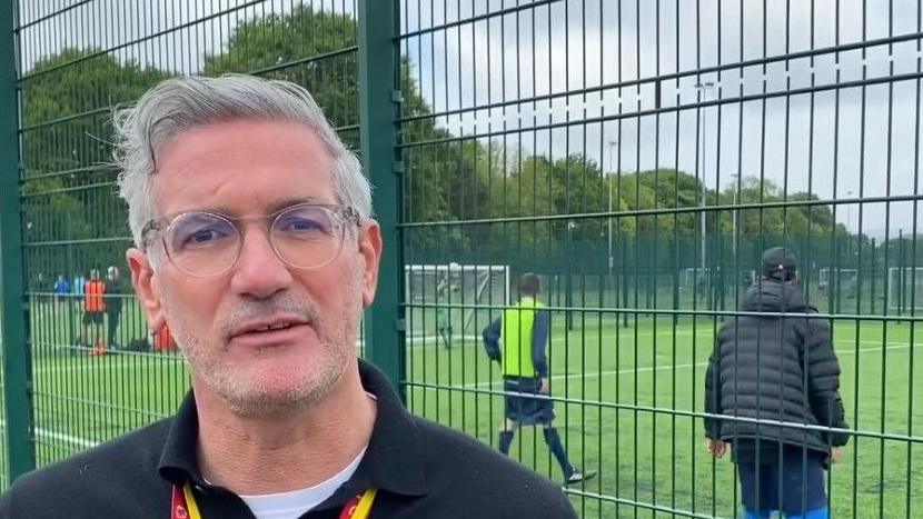 Craig McManus, Acting Head of MFC Foundation, is looking at the camera. He has short grey hair and is wearing a black jumper, yellow and red lanyard and glasses. Players are walking at the football pitch behind him.