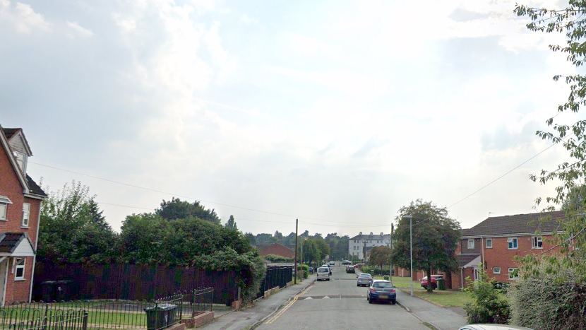 A residential street has houses and flats on either side and cars parked on both sides of the street
