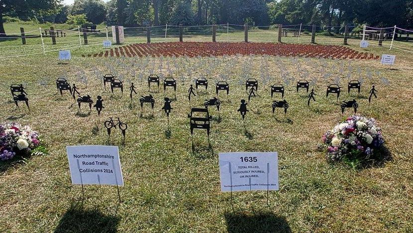 Black silhouettes representing people, cars and bicycles planted into grass. There are notices planted in front showing 1635 - the number of people killed or injured on the county's roads in 2024. There are bouquets of flowers on the left and right. There are grey dove silhouettes behind and small red county flags behind those.