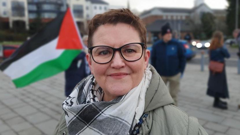 Catherine Hutton smiles as she stands in front of a group of people. One person is holding a Palestine flag. Catherine has black glasses and short brown hair. She is wearing a black and white palestinian keffiyeh.