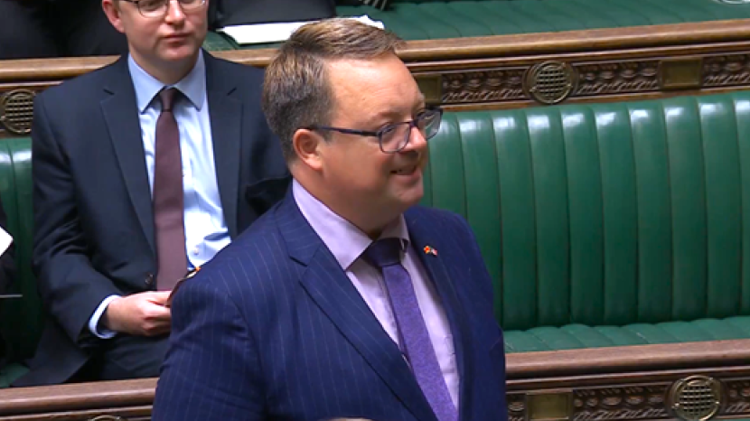 A man with short brown hair and glasses, wearing a navy pintstripe suit with lilac shirt and a purple tie. His body and face are angled away from the camera and he is speaking. In the background a man in a dark suit and maroon tie can be seen sitting on a row of green cushioned benches with ornate wooden carvings at the top.