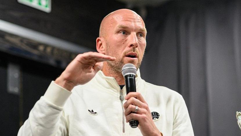 Newcastle United goalkeeper John Ruddy speaking into a microphone while seated on stage. He has a short brown beard and is bald. He is wearing a white jumper decorated with the club's logo.