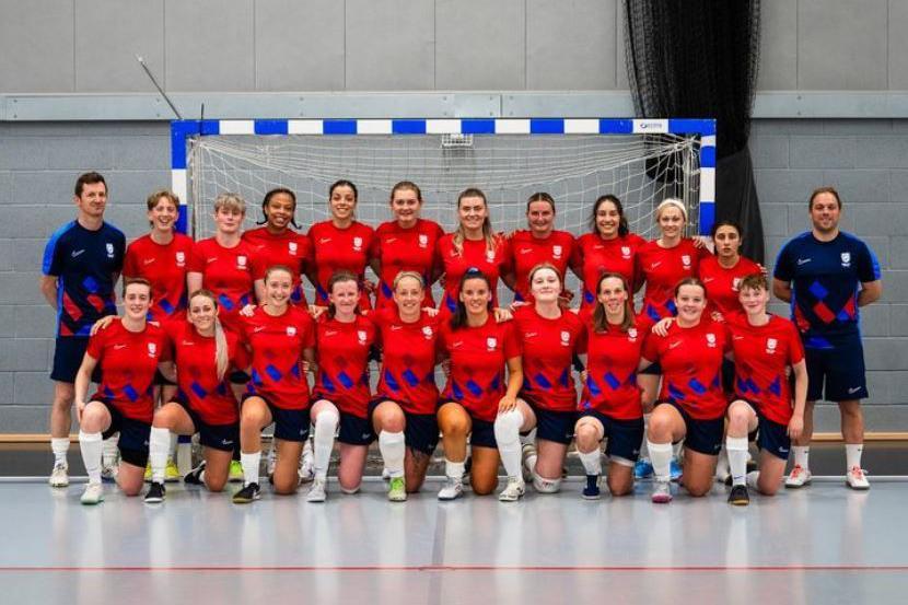 The high performance group, made up of 20 players in red shirts and black shorts, accompanied by two coaches in blue shirts and dark shorts. They are standing in front of a futsal goal, inside a large sports building with hard floors.