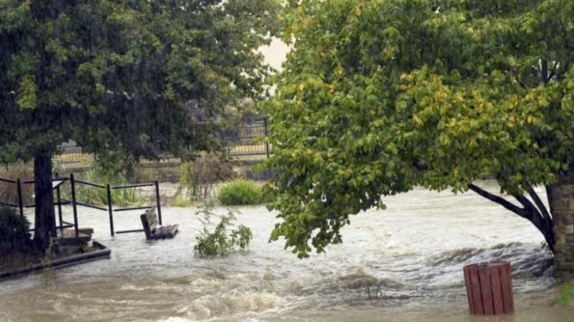 A fast-flowing flood in an urban park area - a litter bin is half-way up with flooding, the water right up to the sea of a bench and the floor cannot be seen anywhere.