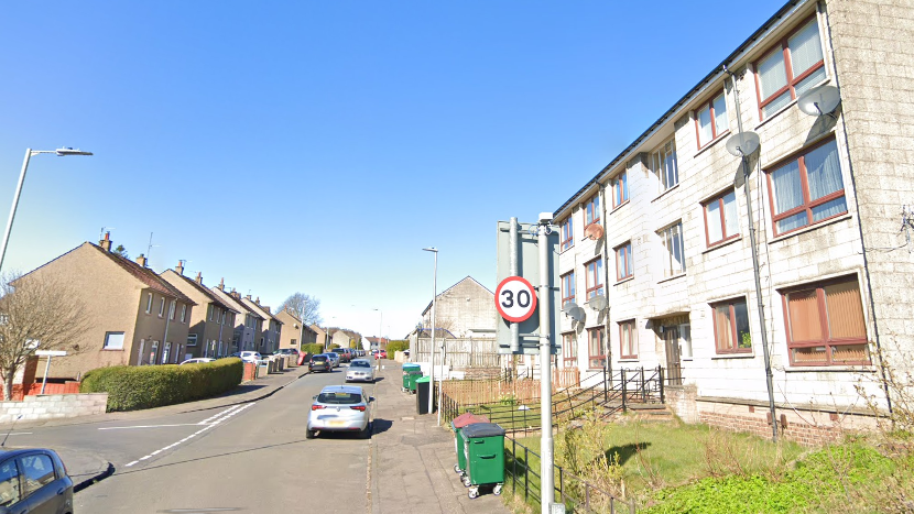 Terraced housing and flats line the street. There are two large green bins. It is a sunny day and the sky is blue. There are cars parked along the street.