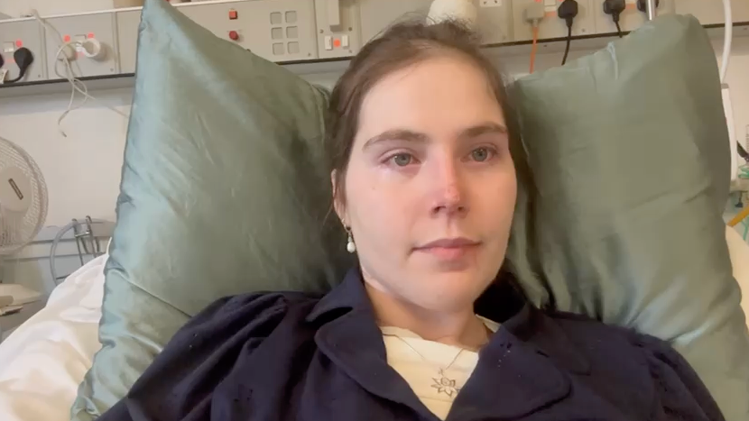 Lily McGarry lying in a hospital bed. They are wearing a dark garment and resting on two pillows. The background includes typical hospital equipment and electrical outlets.

