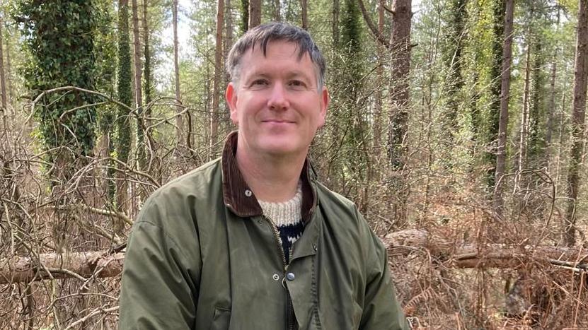 A man with short grey hair stands in a wooded area and smiles at the camera. He is wearing a green coat with a black and white wool top underneath.