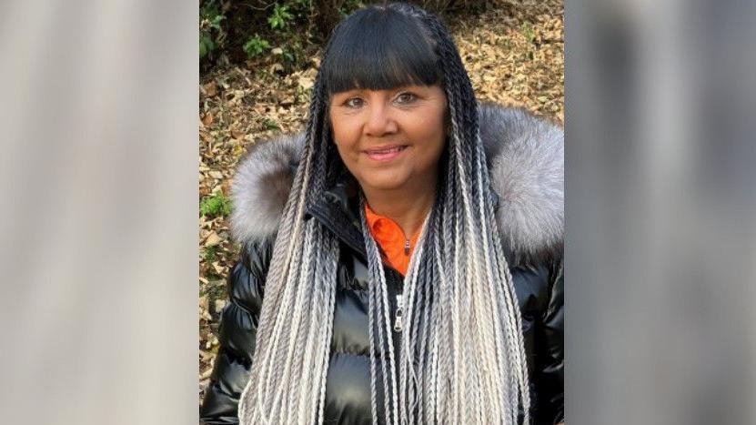 Dot Taylor is smiling at the camera. She has a long braided hair, which is black in the roots and died white in the ends. She has a fringe and is wearing a black puffer jacker with fluffy hood. Her top is orange. 