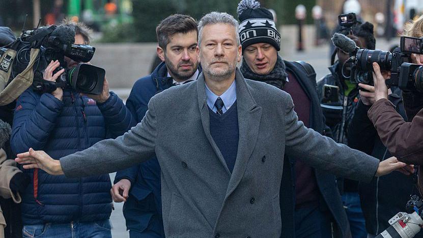 Nathan Gill, wearing a grey coat, blue tie and blue shirt, with his hands held out, holding back a group of journalists and camera crew.