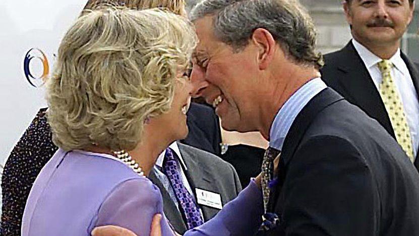 Camilla Parker-Bowles (as was) and then-Prince Charles leaning in for a kiss. Camilla is wearing a purple dress and a pearl necklace and Charles a dark suit and blue shirt. Both are smiling.