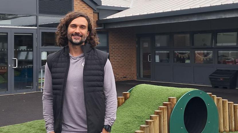 Image of a man with dark curly hair and a beard.  He is smiling at the camera and is wearing a black body warmer, a grey top and black sports watch.  In the background can be seen a school playground with a tunnel and fake grass and a number of classrooms.