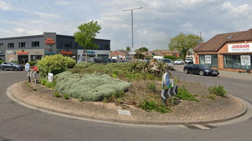 A roundabout with green bushes in the centre at the junction of two routes. There are several cars on the roundabout and at the junctions. Two shops are also visible on the junction with houses in the background.