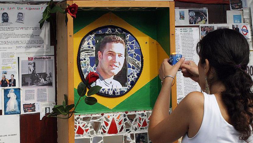 A woman with her back to the camera lights a candle in front of a shrine which features a photo of Jean Charles at the centre of a painted depiction of the Brazilian flag. 