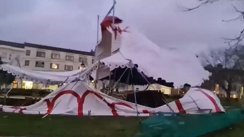 Torn big top tent with red markings in a park with trees behind.