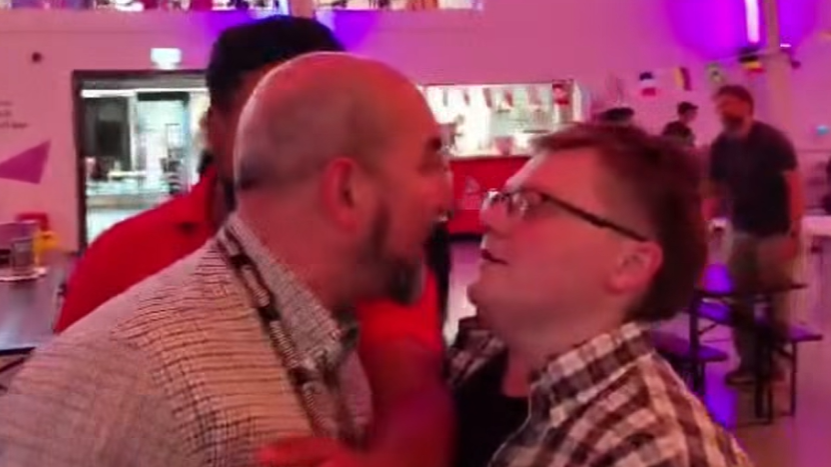 A man with a bald head wearing a checked shirt has an argument with a person in a checked shirt, glasses and short brown hair
