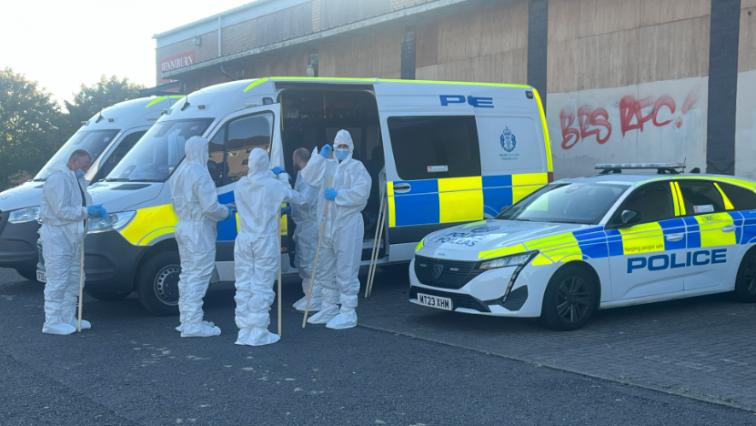 Five forensic officers in white overalls stand next to two police vans and a police care in a car park with a business premises behind them