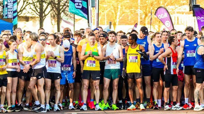 Runners wearing vests and shorts line up on the start line of a race.