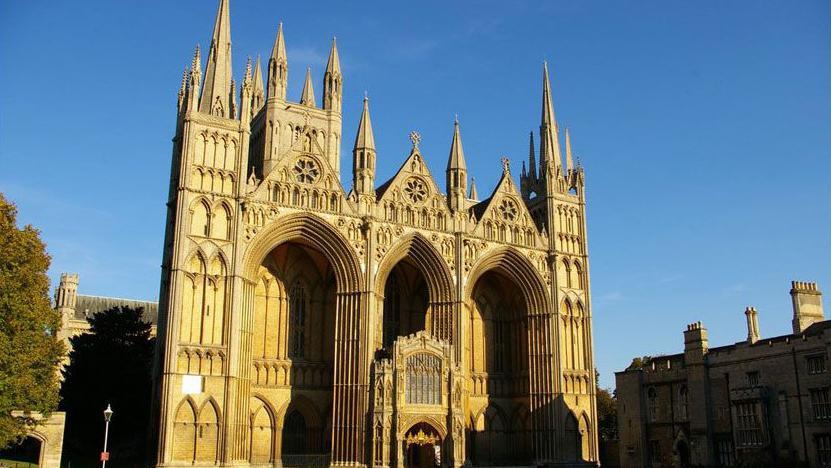 The west end of Peterborough Cathedral. It has three huge arches, and many pinnacles rising above it. It is made of golden-coloured stone. Above it is blue sky. In front is the green grass of a cathedral close.