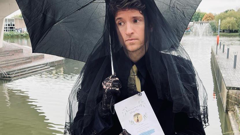 Greg James before the ceremony, dressed in black and holding the order of service. He wears a veil and lace gloves and holds an umbrella