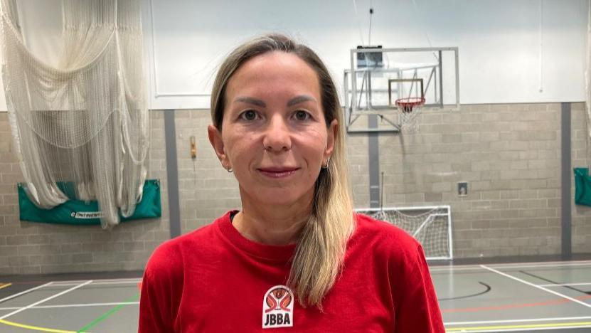 Timea Kraft, with light brown hair pulled back in a loose ponytail stands in a gymnasium. She is wearing a red t-shirt with the letters "JERSEY" in white on the front, and a circular logo above it that says "JBBA". She is looking directly at the camera with a slight smile. Behind her is a basketball hoop. The walls of the gym are made of gray cinder blocks. Green curtains are visible on the left and right sides of the image. The floor has a variety of court lines painted on it.