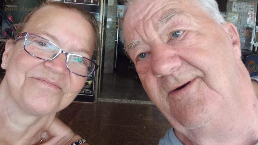 A selfie picture of Julie Green, 60, who has brown hair tied back and is wearing purple rimmed glasses, with Leslie Green, with grey hair and a grey t-shirt.