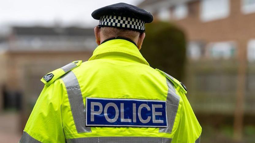 The back of a police officer, in a high-visibility neon yellow jacket with the word police on the back, is shown